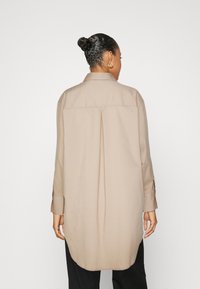 Camicia beige a maniche lunghe con colletto, caratterizzata da una plissettatura sul retro e un orlo curvo, realizzata in tessuto morbido e leggero.