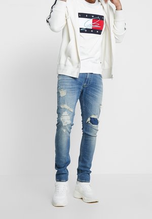 Felpa con cappuccio bianca con zip sopra una t-shirt bianca con grafica. Indossata con jeans blu strappati e sneakers bianche. Outfit casual con strappi visibili nei jeans.