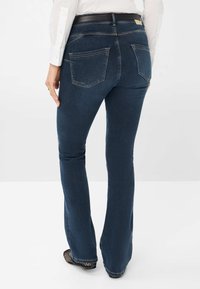 Jean évasé bleu foncé à la texture lisse, avec deux poches arrière et une taille noire. Porté avec une chemise blanche et des chaussures à motifs.