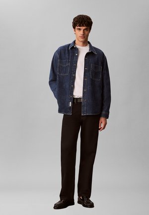 Mann trägt dunkelblaue Jeansjacke über weißem Hemd, schwarze Hose und schwarze Schuhe, steht vor grauem Hintergrund.