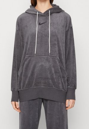 Osoba nosząca luźną ciemnoszara bluzę z kapturem Nike z białymi troczkami i pasujące spodnie na jednolitym tle.