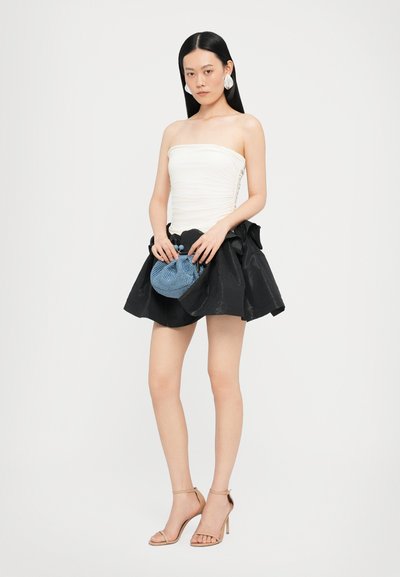 Top blanco sin tirantes con textura fruncida, combinado con una falda negra acampanada, sosteniendo un pequeño bolso azul de tejido; unos tacones beige completan el look.