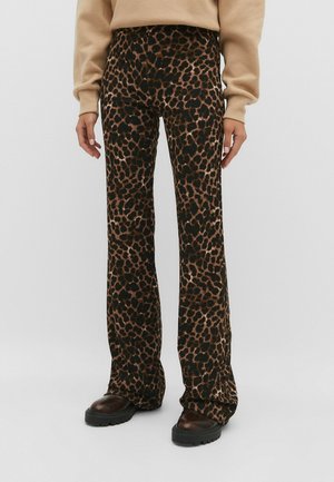 Personne portant un pantalon flare imprimé léopard marron et noir, un haut beige à manches longues, et des chaussures épaisses marron foncé sur un fond uni.