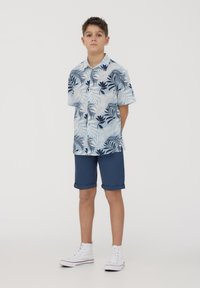 Camicia azzurra a maniche corte con motivi di foglie tropicali scuri, abbinata a pantaloni corti con orlo arrotolato di colore navy e sneakers alte bianche.