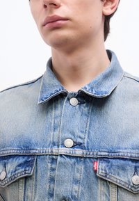 Νεαρό άτομο που φοράει ένα ανοιχτό γαλάζιο τζιν μπουφάν με ασημί κουμπιά και κόκκινη ετικέτα με το λογότυπο Levi's στην τσέπη στο στήθος.