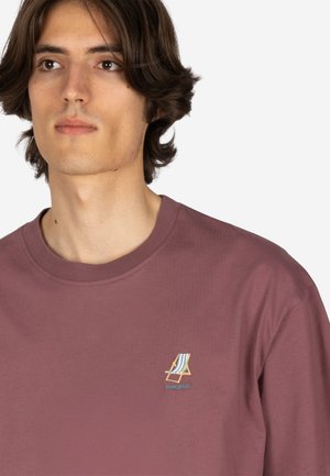 Junger Mann mit mittelbraunen Haaren, der ein mauvefarbenes T-Shirt mit einem kleinen gestickten Strandstuhl und dem Schriftzug "HOME OFFICE" trägt.