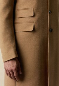 Kamelwollmantel mit weicher Textur, ausgestattet mit zwei Klappentaschen, einem Knopfverschluss und tailliertem Schnitt.
