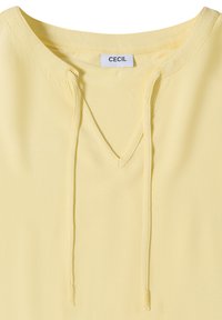 Haut jaune col en V en tissu texturé avec cordons coulissants à l'encolure, avec une étiquette de la marque CECIL à l’intérieur du col.