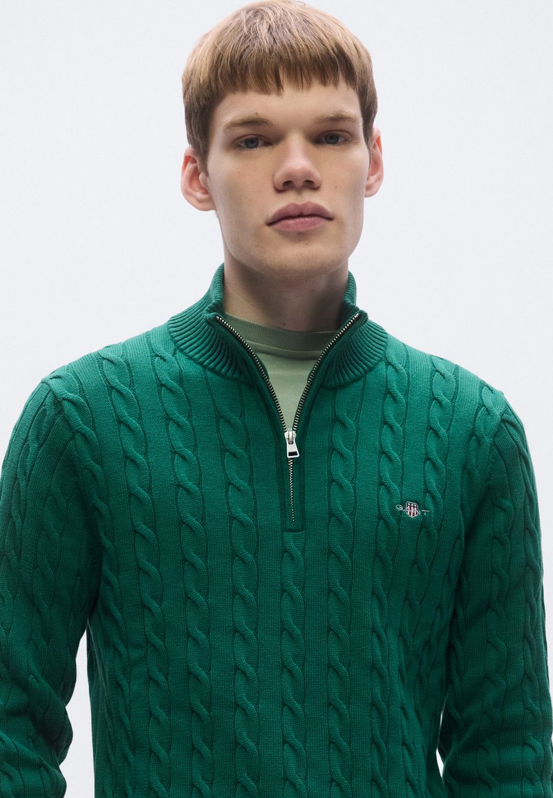 Green knitted sweater with a cable pattern, zippered collar, and a small embroidered logo on the chest. Texture appears soft and thick.