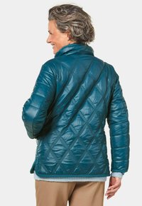 Türkisfarbene Steppjacke mit diagonalen Rautenmustern, hohem Kragen und strukturierten Akzenten. Besitzt eine lockere Passform und lange Ärmel.