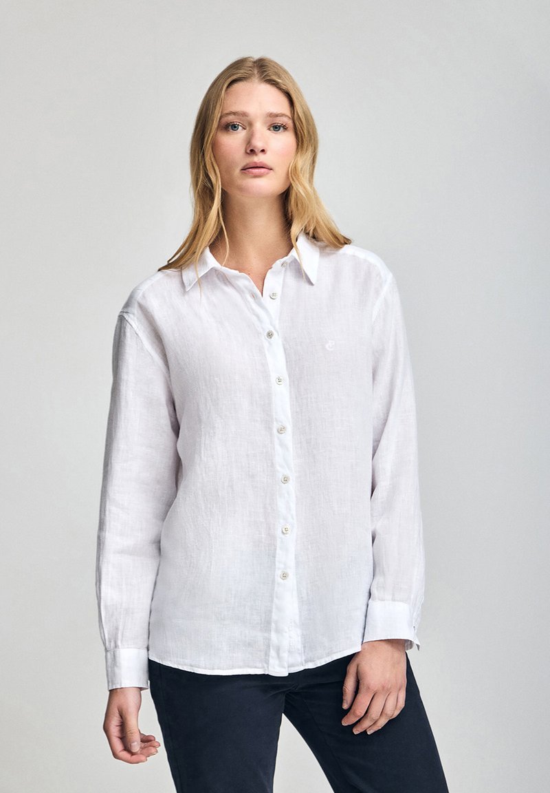 Chemise blanche en lin à manches longues avec une fermeture à boutons sur le devant, col pointu et poignets, présentant une texture subtile et un petit logo sur la poitrine.