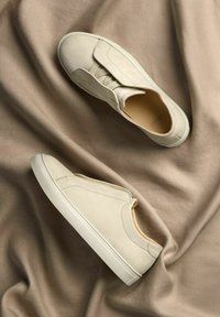 Beige slip-on shoes made of textured leather with elastic panels, featuring a white rubber sole and minimalist design, set on a soft brown fabric.