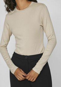 Beige langermet topp laget av mykt stoff, med rund hals og tettsittende design, kombinert med høyhårede svarte bukser.