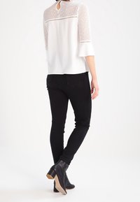 Blusa bianca con pannello in pizzo e dettaglio della schiena a chiave, abbinata a jeans neri attillati e stivaletti con tacco marrone chiaro.