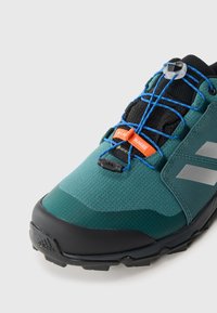 Chaussure de sport verte et noire avec une tige texturée, des lacets orange, un système de bungee et une semelle noire robuste avec le logo Adidas.