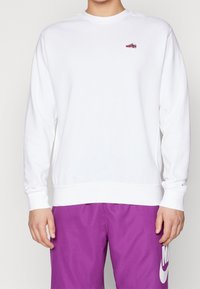 Vit sweatshirt med långa ärmar, ribbade muddarna och en liten röd och grå grafisk patch. Bärs över lila shorts, vilket framhäver färgkontrasten.