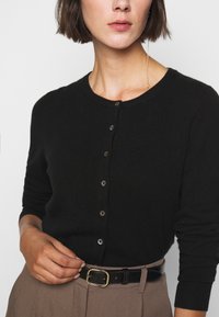 Cardigan nero a maglia con scollatura rotonda, dotato di chiusura con bottoni e dettagli a coste sui polsini, indossato su pantaloni beige con una cintura nera.