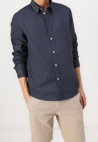 Camicia button-up in denim navy con maniche lunghe, bottoni bianchi e vestibilità rilassata, abbinata a pantaloni beige. Texture morbida, design semplice.