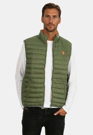Man with light beard wearing green quilted sleeveless vest over white long-sleeve shirt, black pants, hand in pocket, neutral background.