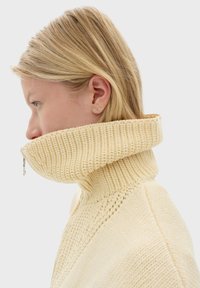 Cremfarbener Strickpullover mit hohem geripptem Kragen, der einen halben Reißverschluss und ein strukturiertes Muster entlang des Körpers aufweist.