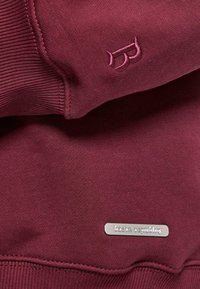 Bordeauxfarbenes Sweatshirt mit gerippten Bündchen, das ein dezentes gesticktes Logo sowie ein metallisches Etikett mit der Aufschrift "basix republic" präsentiert. Weicher Stoff.