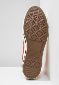 Red canvas sneaker with a rubber sole. Features a textured brown bottom, a cream side stripe, and a star logo. Design includes geometric patterns.