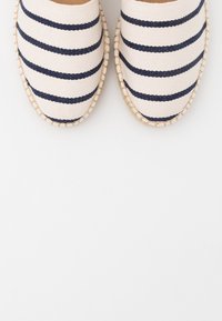 Espadrilky s námořnickými modrými vodorovnými pruhy na béžové tkanině a tkanou juteovou podrážkou zobrazené z vrchu na bílém pozadí.