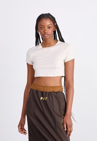 Kreminė trumpomis rankovėmis, trumpa T-marškinėliai su apvaliu kaklu ir Nike logotipu, derinami su rudomis kelnėmis, turinčiomis elastinį juosmenį ir prekės ženklo akcentus.