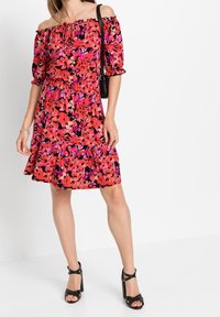 Červené a růžové květinové šaty s odhalenými rameny, krátkými nafouklými rukávy a volánkovým lemem, doplněné černými sandály na podpatku. Černá kabelka jako doplněk.