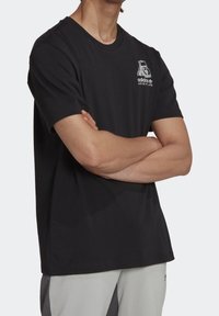 Man med svart Adidas Adventure t-shirt med en björnlogotyp på bröstet, armarna i kors, i kombination med ljusgrå byxor.
