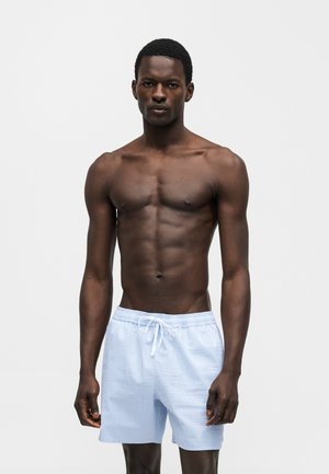 Homme torse nu à la peau foncée portant un short à cordon rayé bleu clair et blanc, debout devant un fond gris clair uni.