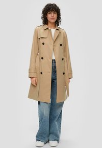 Manteau beige en tissu léger avec un design à double boutonnage, boutons noirs et poches latérales, associé à un pantalon en jean à jambes larges.