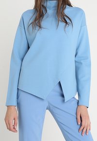 Vaaleansininen, pitkähihainen paita, jossa on korkea kaulus ja epäsymmetrinen, kulmikas helma. Valmistettu sileästä, puolistrukturoidusta kankaasta.