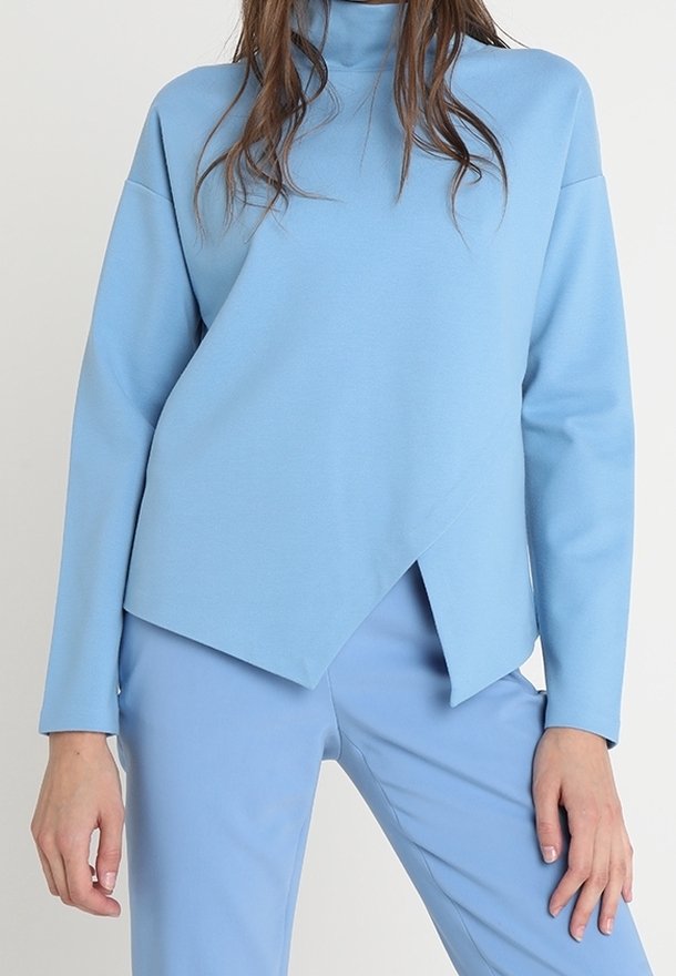 Vaaleansininen, pitkähihainen paita, jossa on korkea kaulus ja epäsymmetrinen, kulmikas helma. Valmistettu sileästä, puolistrukturoidusta kankaasta.