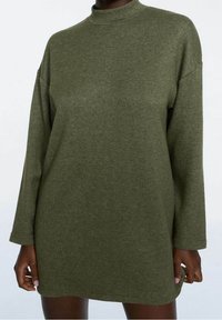 Henkilö, joka on pukeutunut suuriin oliivinvihreään pitkähiharaiseen neulemekkoon, jossa on korkea kaula-aukko, kädet rentoina sivuilla.