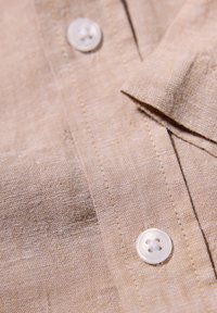 Beige linne tyg med en mjuk, texturerad yta pryds av en vit knapp med fyra hål och fint avslutade sömmar längs kanten.