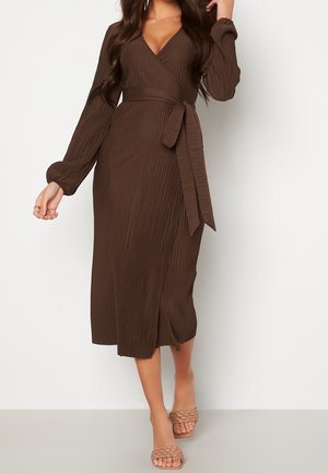 Femme portant une robe portefeuille marron à manches longues et plissée, avec une ceinture nouée à la taille et des sandales plates tressées beiges, marchant devant un fond uni.