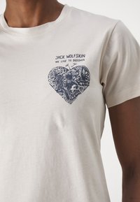 Lichtbeige katoenen t-shirt met een donkerblauw hartontwerp en natuurelementen, met de tekst "JACK WOLFSKIN WE LIVE TO DISCOVER" erboven.
