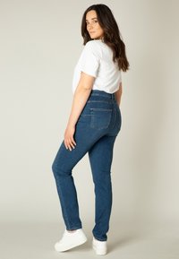 Blauwe high-waisted jeans met een slim fit, voorzien van twee achterzakken en gestikte naden, gecombineerd met een witte T-shirt met korte mouwen en witte sneakers.