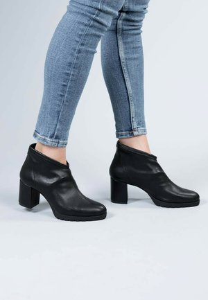 Botas de cuero negro hasta el tobillo con un acabado suave, que cuentan con un tacón cuadrado alto y un diseño elegante y minimalista. Combinadas con jeans ajustados azules.