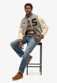 Grijze en crème varsityjack met borduursel, geruit overhemd, blauwe jeans en bruine veterschoenen, zittend op een zwarte kruk tegen een witte achtergrond.