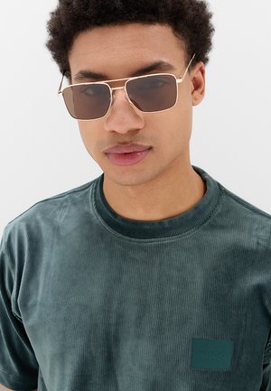 Giovane uomo con occhiali da sole rettangolari con montatura dorata e maglietta in velluto a coste verde scuro con piccolo logo sul petto.
