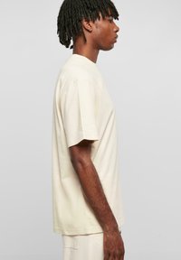 Tricou bej cu mânecă scurtă, cu o croială lejeră, fabricat din bumbac, având guler rotund și un design simplu. Textura materialului este vizibilă.