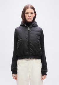 Γυναίκα που φοράει μαύρο γιλέκο με φερμουάρ και κουκούλα πάνω από μαύρο μακρυμάνικο μπλουζάκι και εκρού παντελόνι, στέκεται μπροστά σε ένα απλό ανοιχτό φόντο.