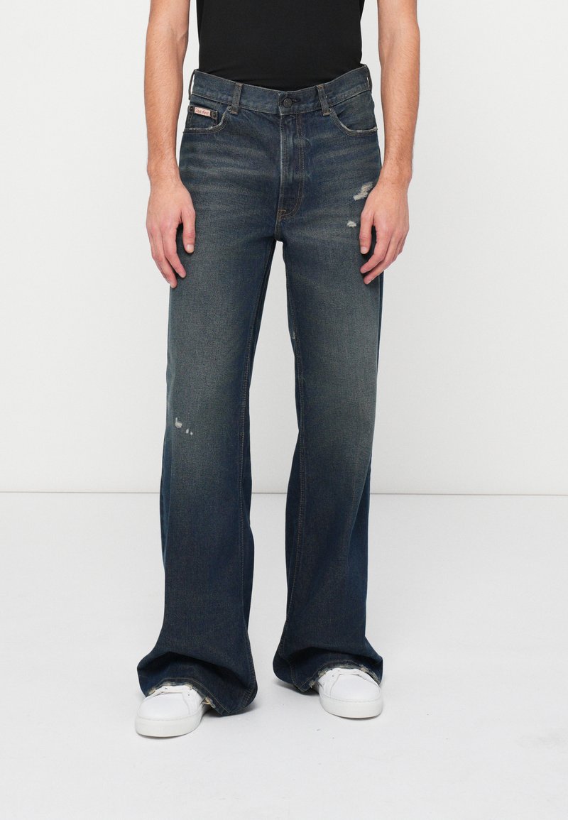 Jeans largi de culoare albastru închis cu detalii uzate, confecționați din denim. Au un design clasic cu cinci buzunare și închidere cu nasture. Adidași albi.