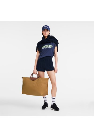 Braune, große Tragetasche mit kurzen Ledergriffen; Modell in einem marineblauen T-Shirt mit grünem Grafikdesign, marineblauen Shorts, schwarzen Sneakers, weißen gestreiften Socken und einer blauen Kappe.