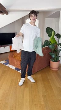 Dvé košele svetlobéžovej a bledozelenej farby, ktoré drží osoba v bielej košeli a čiernych vzorovaných nohaviciach. Interiér s rastlinami a nábytkom.