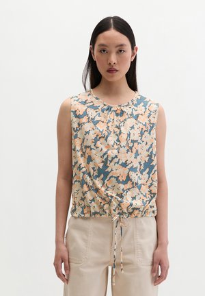 Femme aux longs cheveux foncés portant un haut fleuri sans manches avec un ourlet ajustable par cordon et un pantalon beige clair, debout sur un fond blanc.