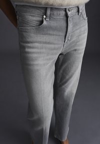 Jeans grigi in denim indossati da una persona con le mani nelle tasche, che mostrano le cuciture frontali e laterali su uno sfondo scuro.