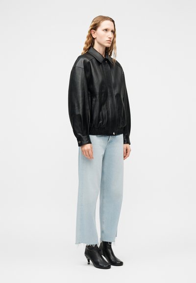 Γυναίκα που φοράει μαύρο δερμάτινο μπουφάν, ανοιχτό μπλε τζιν με φαρδιά πόδια και ξεφτισμένες άκρες, και μαύρα μποτάκια αστραγάλου μπροστά σε απλό γκρι φόντο.